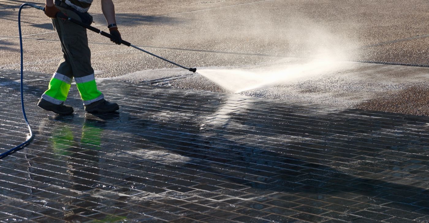 Worker in high-visibility vest pressure washing a paved surface with water spray